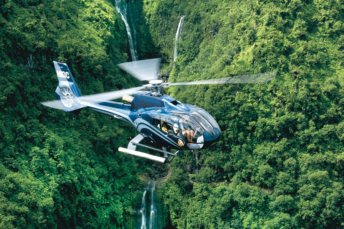 Blue Hawaiian helicopter flying past lush green cliffs and waterfalls over Maui’s coastline.